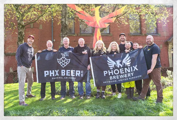 Team members from the two partnered breweries standing outdoors on the grass in front of a red-brick building, proudly holding banners for PHX Beer Co. (Arizona) and Phoenix Brewery (UK) with a large phoenix sculpture behind them.